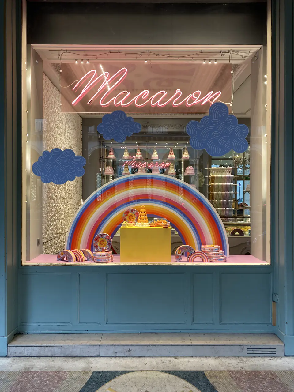 Vitrine de pâtisserie Ladurée : nuages bleus en adhésif découpé sur le verre, arc-en-ciel et pyramide de macarons installés derrière la vitre