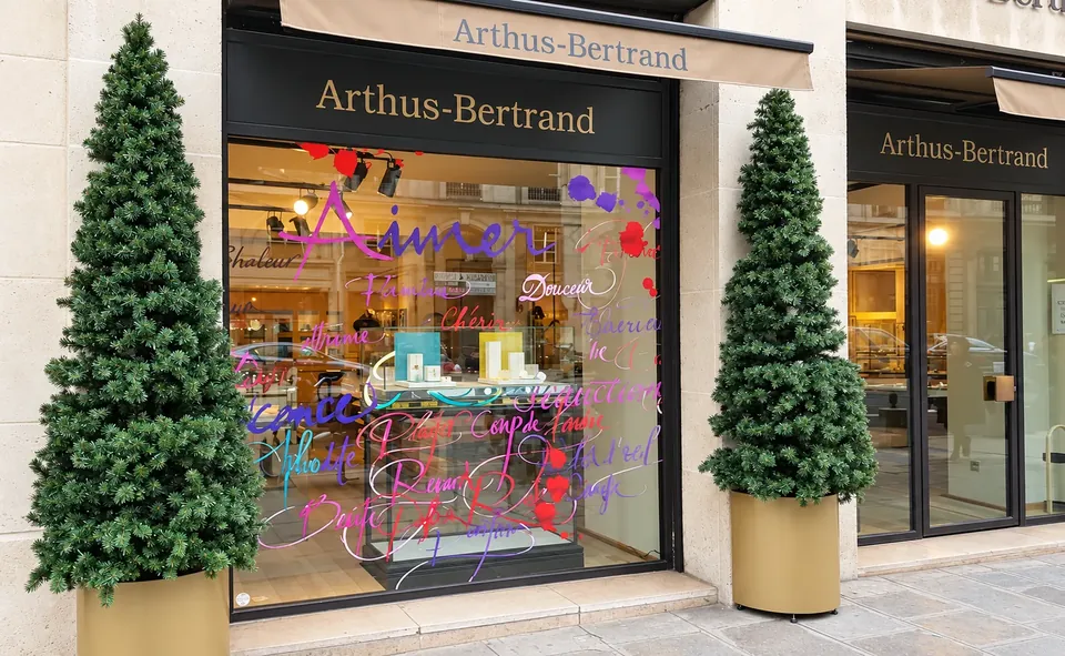 Vitrine de Noël Arthus-Bertrand : lettrage manuscrit multicolore en vitrophanie (« Aimer », « Chaleur », « Douceur ») sur la vitrine, encadrée de deux sapins en pot doré