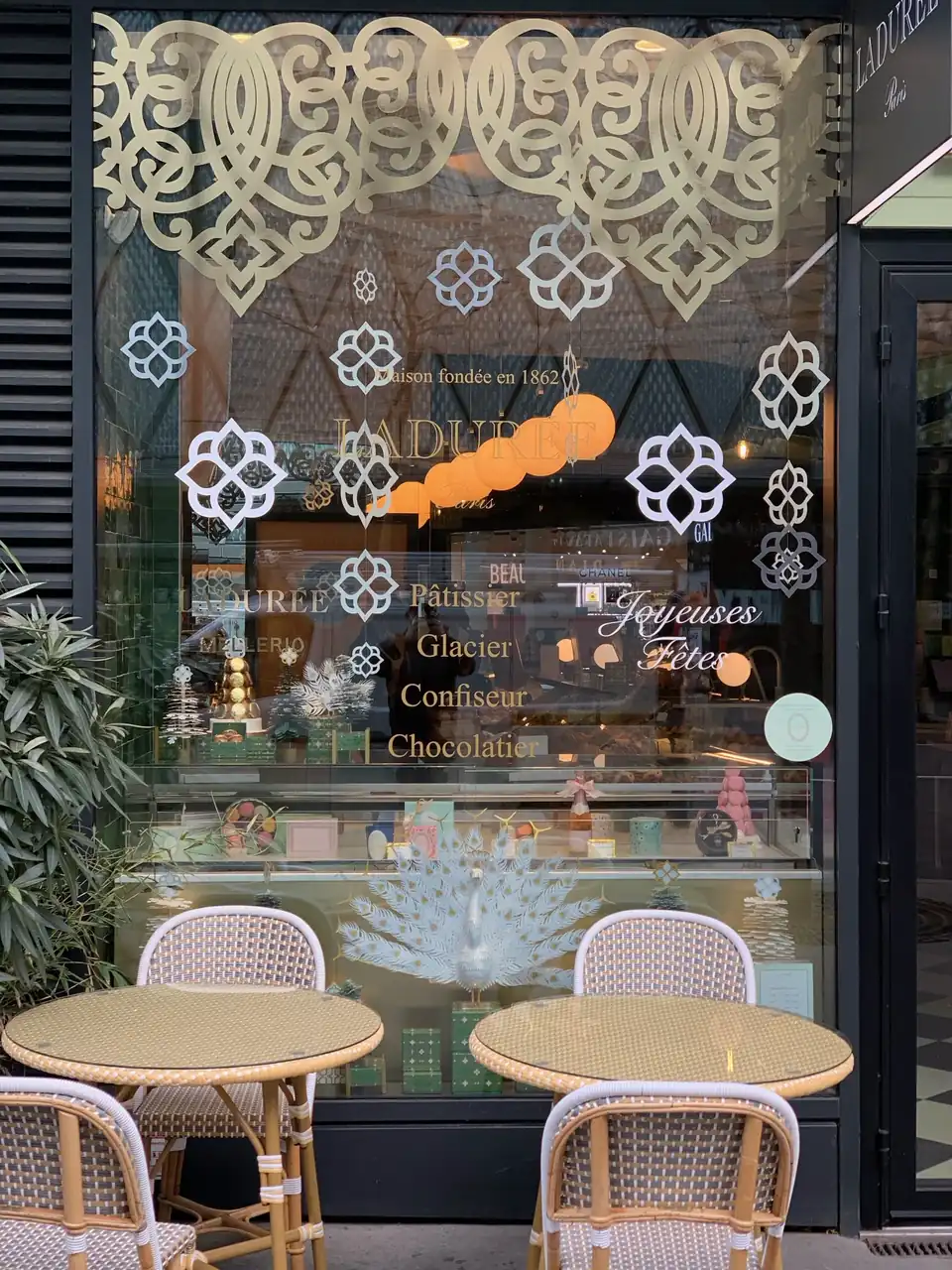 Vitrine de Noël Ladurée : arabesques dorées en partie haute, rosaces blanches et lettrage « Joyeuses Fêtes » en vitrophanie au-dessus de la terrasse