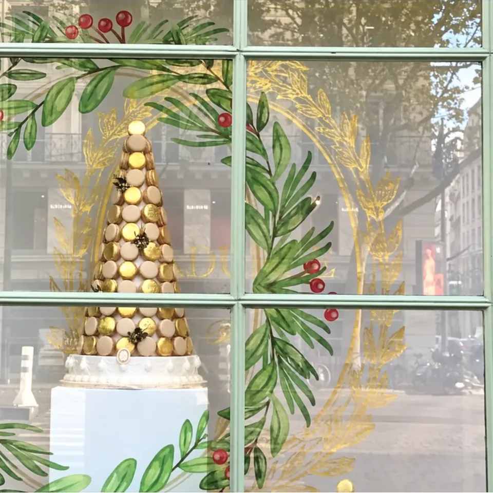 Vitrine de Noël Ladurée vue de près : couronne de feuillage vert, baies rouges et épis dorés en vitrophanie autour d'une pyramide de macarons