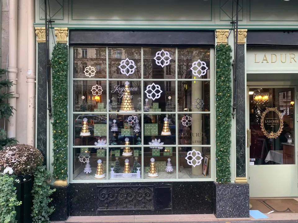 Vitrine de Noël Ladurée : rosaces blanches et lettrage « Joyeuses Fêtes » en vitrophanie sur les vitrages à petits bois, pyramides de macarons dorées derrière le verre