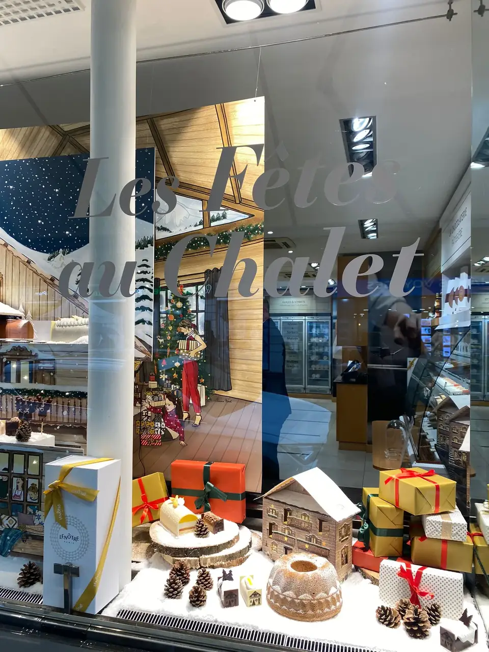 Vitrine de Noël Lenôtre : grand lettrage « Les Fêtes au Chalet » sur le vitrage, décor de chalet enneigé et paquets cadeaux en vitrine