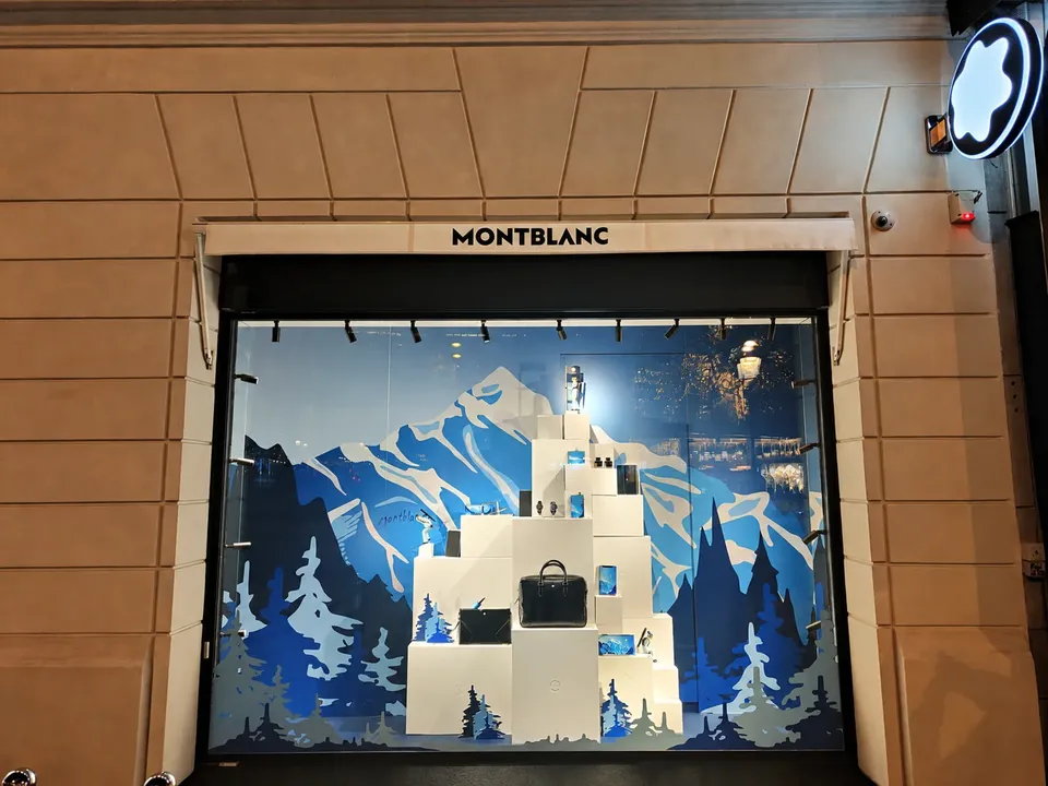 Vitrine de Noël Montblanc : décor de montagnes enneigées et de sapins bleus en fond de vitrine, maroquinerie exposée sur des cubes blancs au premier plan